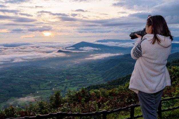 Turista de la mujer está utilizando una cámara DSLR fotografiando el paisaje de la naturaleza la montaña de niebla del sol en el invierno durante el amanecer en el mirador alto en el Parque Nacional de Phu Ruea, provincia de Loei, Tailandia