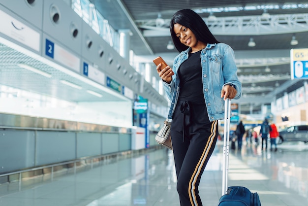 Turista mujer negra con maleta y teléfono esperando la salida en el aeropuerto internacional.