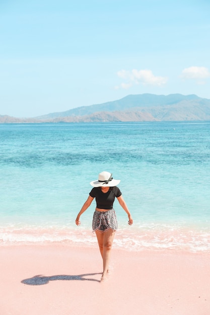 Turista de mujer con sombrero de verano caminando en la playa de arena rosa con vistas al mar y colinas en Labuan Bajo