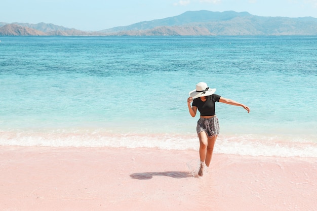 Turista de mujer con sombrero de verano caminando en la playa de arena rosa con vistas al mar y colinas en Labuan Bajo