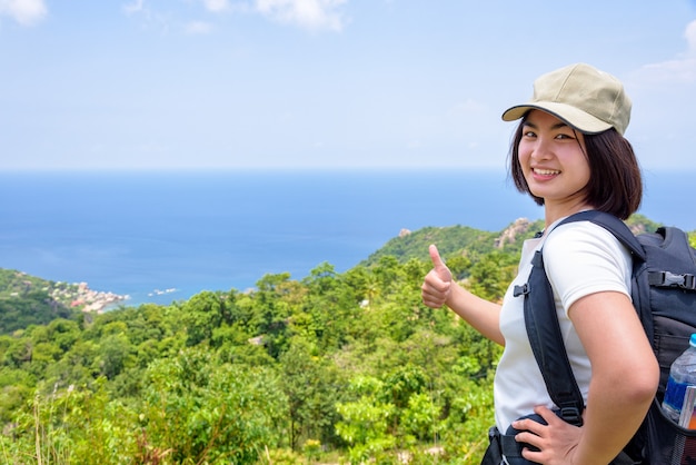 Turista de mujeres con una gorra de mochila levantar el pulgar hacia arriba para el hermoso paisaje natural del mar azul y el cielo desde el mirador escénico alto en Koh Tao, Surat Thani, Tailandia