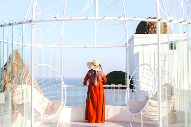 Turista de mujeres en vestido rojo y sombrero de verano mirando al mar desde el balcón superior de vacaciones