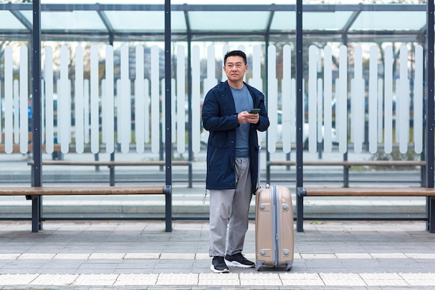 Turista de negocios asiático, esperando un taxi cerca del aeropuerto, usa la aplicación telefónica para alquilar un auto, un hombre con una maleta grande