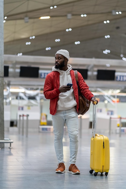 Turista negro esperando para registrarse en los puestos de la terminal del aeropuerto con maleta usando un teléfono inteligente