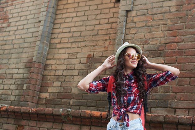 Turista de niña en gafas de sol se encuentra y ajusta su sombrero en la cabeza contra la pared de ladrillo rojo