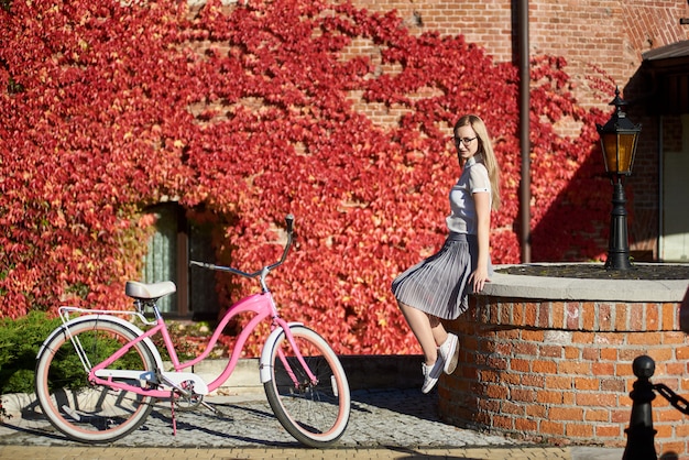 Turista de niña sentada en la pared de ladrillo bajo en bicicleta de dama rosa en brillante cálido día soleado de otoño