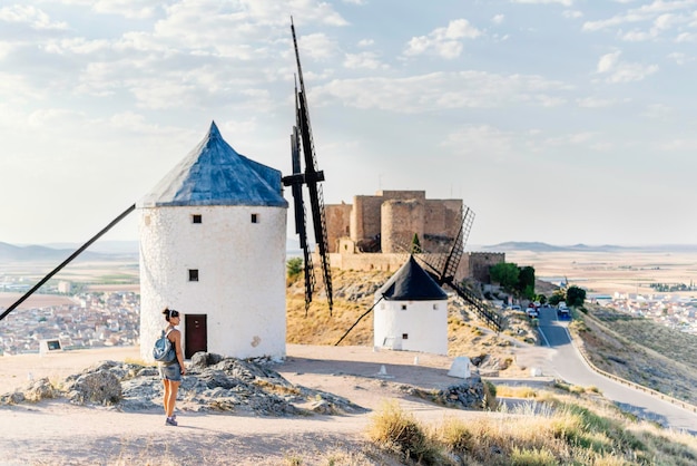 Turista de pie sobre una colina con un grupo de antiguos molinos de viento restaurados y un castillo en Consuegra, Toledo en España