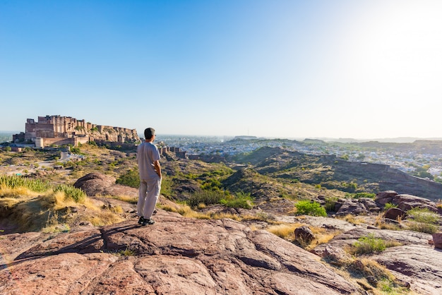 Turista de pie sobre una roca y mirando la vista expansiva del fuerte de Jodhpur desde arriba, encaramado en la parte superior dominando la ciudad azul. Destino de viaje en Rajasthan, India.