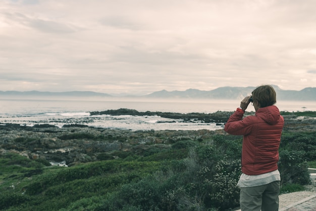 Turista que olha com binocular na linha rochosa da costa em De Kelders, África do Sul, famosa para a observação da baleia.