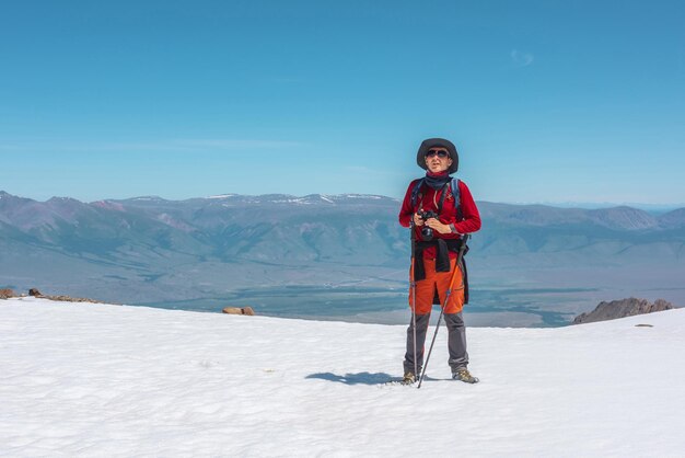 Turista en rojo camina en la montaña nevada cerca del borde del abismo a gran altura bajo el cielo azul en un día soleado Hombre con cámara en la montaña nevada cerca del borde del precipicio con vista a la gran cordillera en la distancia