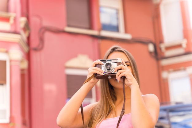 Turista rubia en una calle con casas con fachadas coloridas tomando una foto con la cámara vintage
