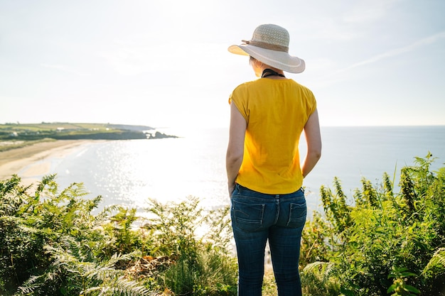 Turista rubio Camiseta amarilla vibrante Sombrero colorido Aventura en la costa del acantilado al mediodía