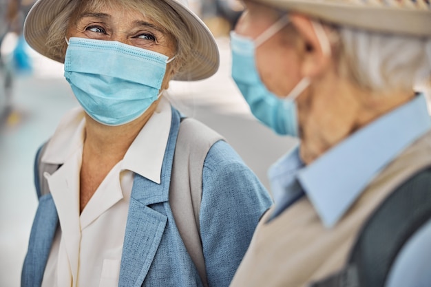 Turista satisfeita com um chapéu e uma máscara facial sorrindo para um homem branco de cabelos grisalhos