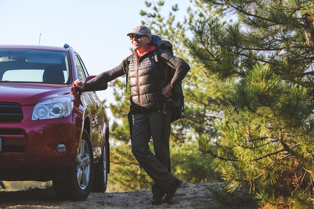 Turista senior feliz con una mochila apoyarse en coche rojo en el bosque