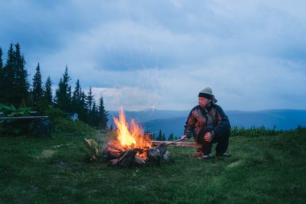 Turista sentado cerca de fuego de campamento en el crepúsculo en la montaña