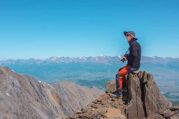 Turista sentado en piedra cerca del borde del abismo a gran altura bajo un cielo azul en un día soleado Hombre con cámara en roca alta cerca del borde del precipicio con maravillosas vistas desde arriba a la gran cordillera de lejos