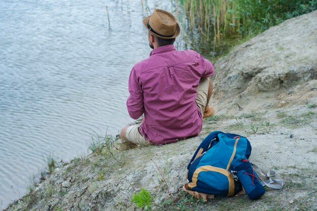 El turista se sienta en la orilla del lago con una mochila para viajar.