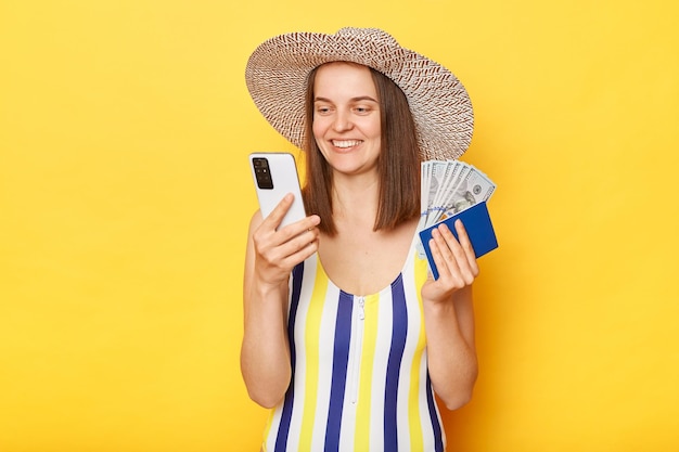 Turista sorridente otimista segurando bilhetes de reserva de passaporte de celular planejando viajar usando maiô listrado posando isolado sobre fundo amarelo