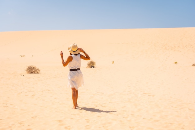 Turista con vestido blanco y sombrero caminando por las dunas del Parque Natural de Corralejo, Fuerteventura, Islas Canarias. España