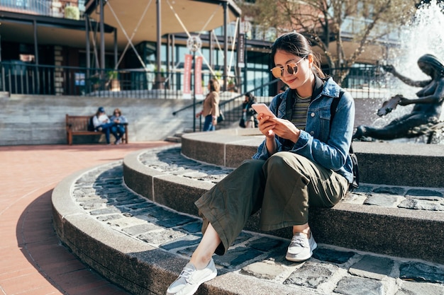 Turistas asiáticas usam mensagens de texto de celular online. jovem segurando smartphone jogar rede social procurando dando direções para Sightseeing spot sentado na fonte em San Francisco