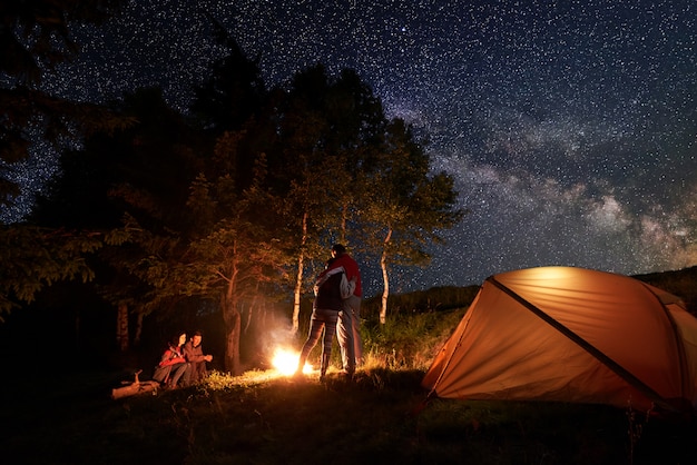 Los turistas durante una noche en el campamento alrededor de una fogata cerca de la carpa naranja. Un par se abraza, el segundo está sentado en un tronco bajo el cielo estrellado y la Vía Láctea sobre un fondo de árboles.