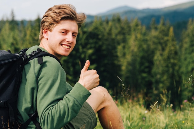 Turistas positivos jovem descansando sentado na grama nas montanhas no fundo de uma bela paisagem olhando para a câmera com um sorriso no rosto e mostrando os polegares