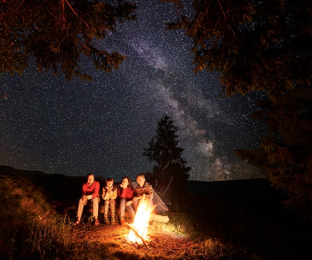 Turistas sentados en un tronco junto a la fogata con una gran llama bajo los abetos bajo el extraordinario cielo cubierto de estrellas.