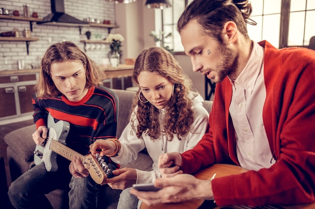 Tutor de enseñanza. Tutor de guitarra talentoso experimentado con un pequeño tatuaje en su rostro enseñando a hermano y hermana