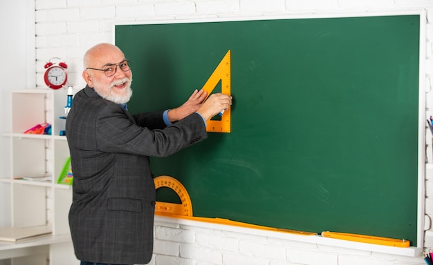Tutor Mann mit Brille zeichnet mit Dreieck auf Tafel zurück zur Schule, aus welchem Winkel Sie aussehen, geometrische Formen, High School, moderne Bildung, älterer Mann, Lehrer, verwenden Triagle beim Zeichnen