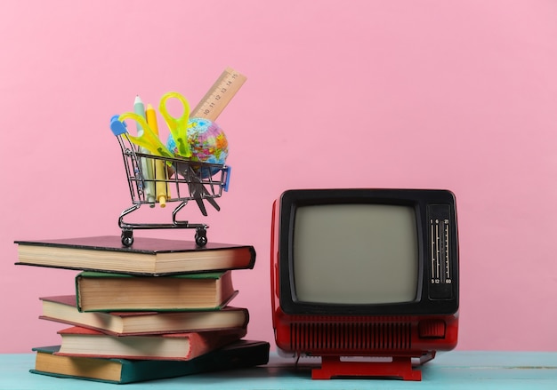 Tv retro y pila de libros, carrito de la compra con útiles escolares sobre fondo rosa. Aprendizaje a distancia por televisión.
