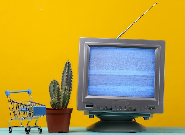 TV-Shop. Antenne altmodischer Retro-Fernseher und Mini-Supermarktwagen mit Kaktus auf Gelb