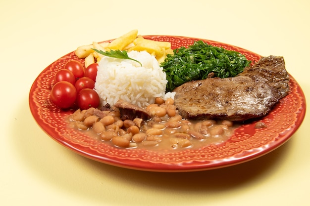 Typisch brasilianisches Grundgericht mit Reis, Bohnen, Steak, Pommes Frites, geschmortem Kohl und Tomate in einem roten Teller im Blick