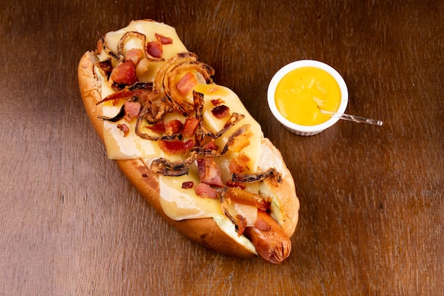 Typischer amerikanischer Gourmet-Hotdog mit Speckschmelzkäse und knusprigen Zwiebeln auf Holztisch, Nahaufnahme mit gelbem Senf