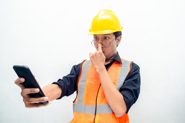 Uau animado trabalhador da construção civil masculino asiático em colete laranja e capacete de segurança amarelo segurando o telefone