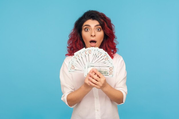 Uau, eu sou rico retrato de mulher surpreendida com cabelo vermelho chique segurando notas de dólar e olhando para a câmera com espanto chocado com a loteria vencedora do estúdio interno tiro isolado no fundo azul