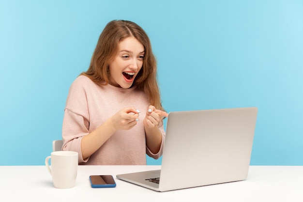 Uau, inacreditável, mulher excitada e surpresa, apontando para a tela do laptop lendo notícias chocantes ou falando em videochamada com expressão espantada, tiro de estúdio interno isolado em fundo azul