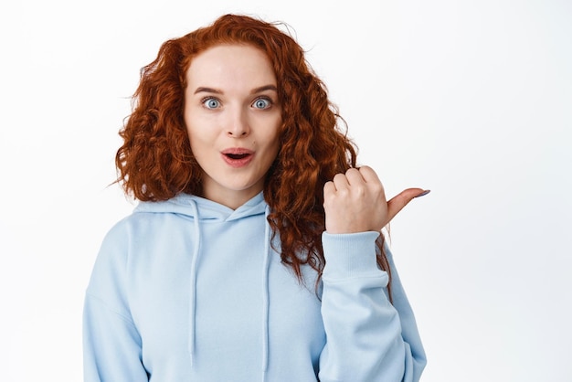 Uau, incrível garota ruiva impressionada com cabelo encaracolado parece animada apontando o dedo para a direita e sorrindo atônita usando capuz fundo branco