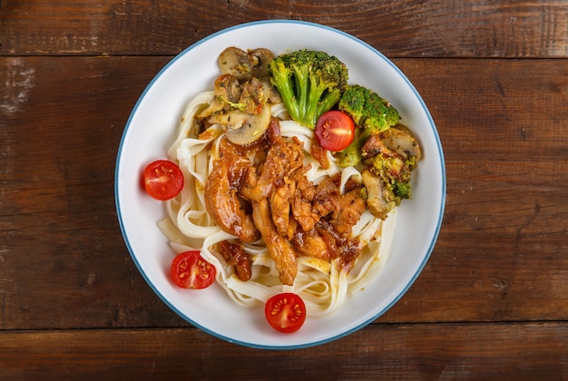 Udon com frango em molho japonês e legumes em um prato sobre uma mesa de madeira. Foto horizontal