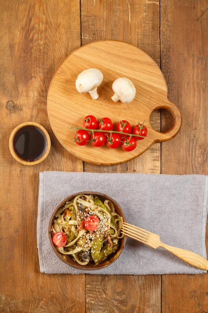 Udon com legumes e cogumelos na mesa em uma placa de madeira ao lado de champignon e tomate e molho de soja em uma prancha