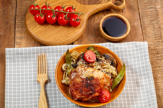 Udon com legumes e frango com sementes de gergelim em uma placa de madeira ao lado de tomates e champignons e molho de soja em uma prancha