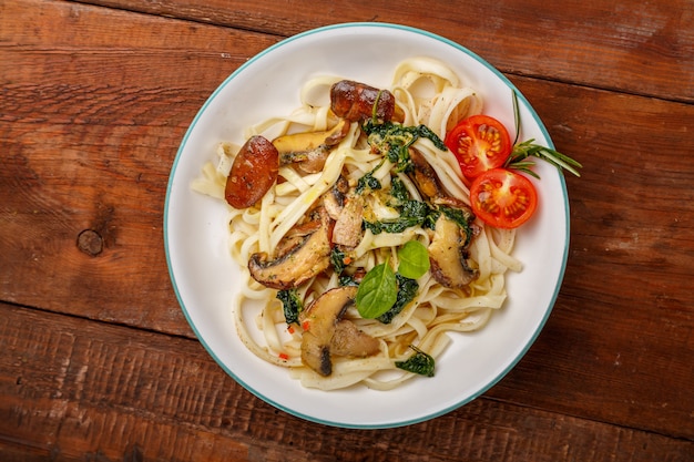 Udon mit Pilzen und Spinat und Kirschtomaten in einem Teller auf einem Holztisch. Horizontales Foto