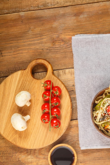 Udon con verduras y champiñones sobre la mesa en un plato de madera junto a champiñones y tomates y salsa de soja en una tabla