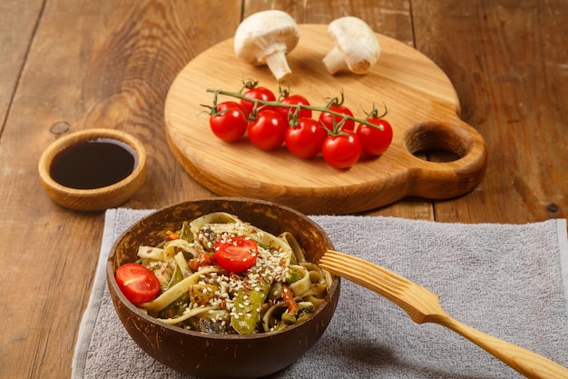 Udon con verduras y champiñones sobre la mesa en un plato de madera junto a champiñones y tomates en una tabla