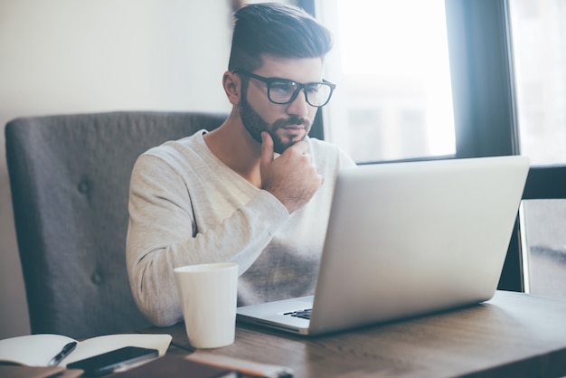 Über Chancen nachdenken. Nachdenklicher junger Mann mit Brille, der am Laptop arbeitet und die Hand am Kinn hält, während er im Büro oder Café sitzt