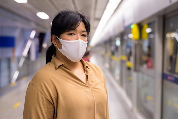 Übergewichtige asiatische frau, die mit maske auf schutz vor ausbruch des koronavirus am u-bahn-bahnhof wartet