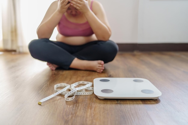 Übergewichtige Frau mit Fettverärgerung gelangweilt von Diäten Gewichtsverlust scheitert Fette Diät und Skala traurige asiatische Frau auf der Waage zu Hause Gewichtskontrolle