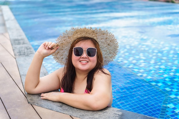 Übergewichtige junge Asiatin trägt orangefarbenen Badeanzug und Strohhut am Strand und entspannt sich im Pool. Glückliche Plus-Size-Frau, fröhlich, lustig, Urlaubsreisen im Sommer