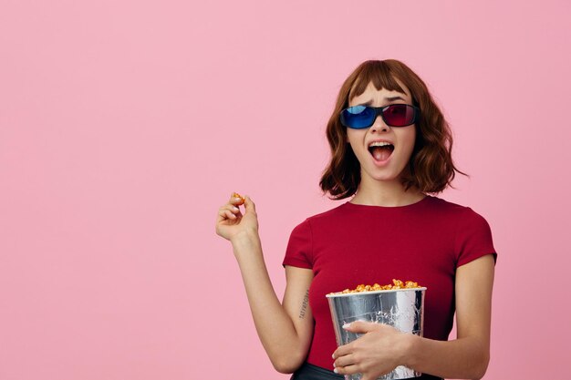 Überglückliche, glückliche, niedliche rothaarige Dame in rotem T-Shirt 3D-Brille mit Popcorn-Film, der isoliert auf über rosa Studiohintergrund posiert Kopierbereich Banner Modekino-Konzept Unterhaltungsangebot
