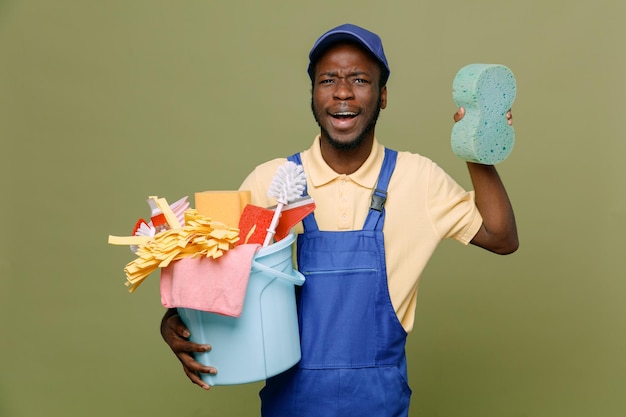 überrascht, Eimer mit Reinigungswerkzeugen mit Schwamm zu halten junger afroamerikanischer Reinigungsmann in Uniform mit Handschuhen isoliert auf grünem Hintergrund