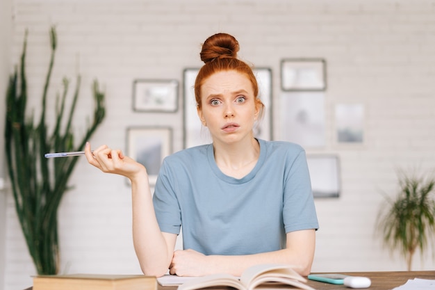 Foto Überrascht enttäuschte rothaarige junge studentin sitzt an einem tisch mit büchern und einem arbeitsbuch